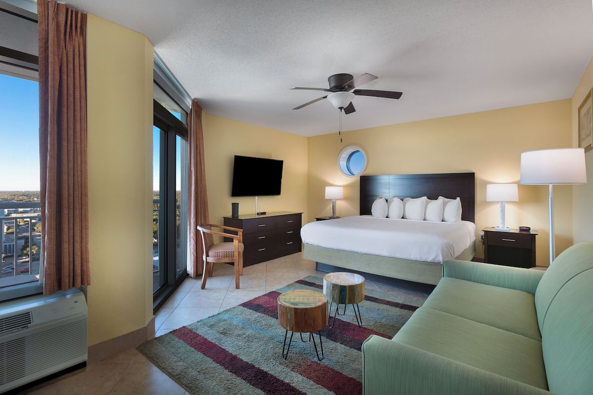 A hotel room with a bed, two bedside lamps, a ceiling fan, a TV, a chair, and a view of the city.