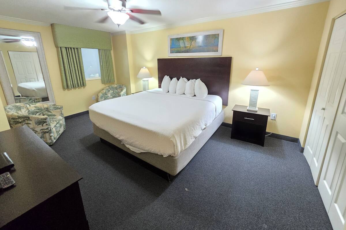 A hotel room with a large bed, a ceiling fan, a desk with a lamp, a chair, and a painting on the wall.
