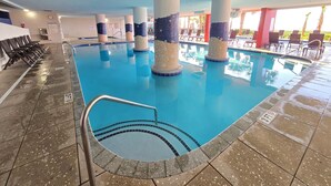 Une piscine intérieure à l’eau claire, entourée d’un sol carrelé et d’un espace aménagé avec des chaises et un parasol.
