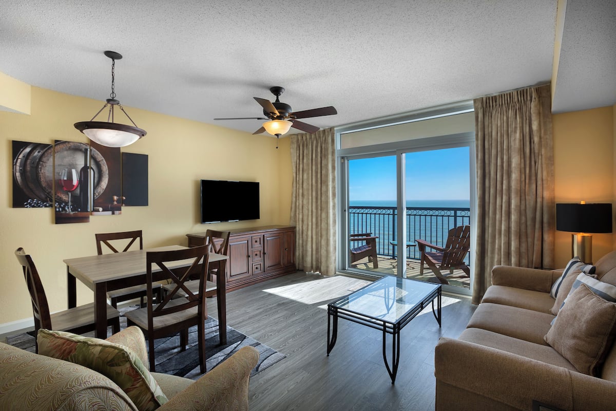 A spacious living area with a balcony, a ceiling fan, a flat-screen TV, and a dining set.