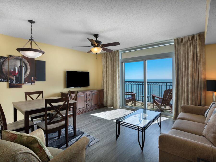 A spacious living area with a balcony, a ceiling fan, a flat-screen TV, and a dining set.