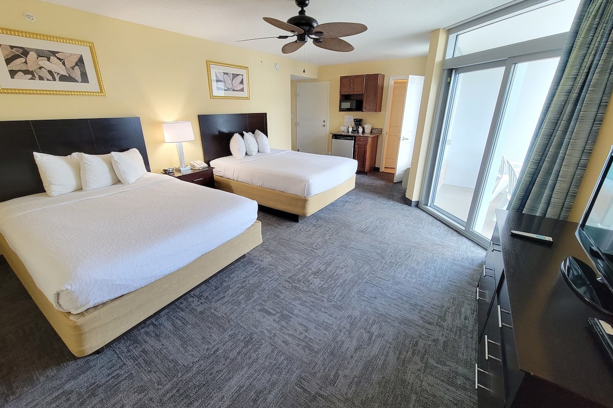 A hotel room with two beds, a ceiling fan, a desk with a TV, and a large window.
