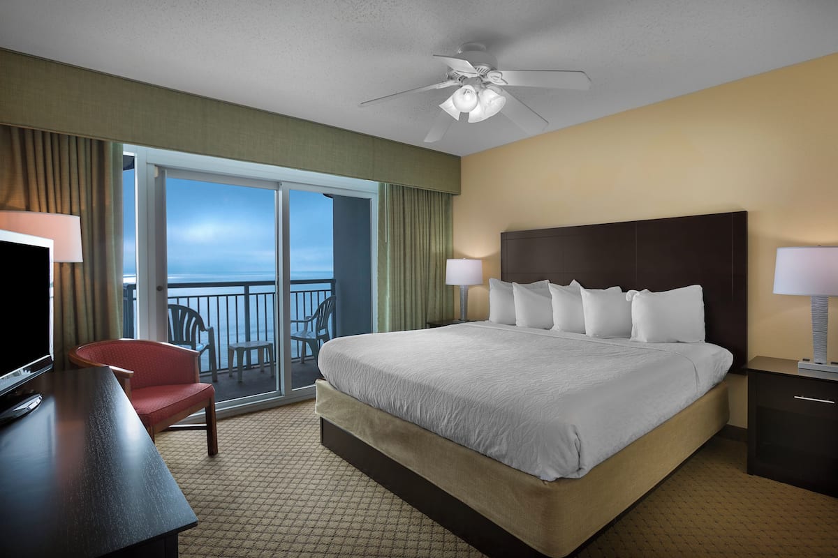 A hotel room with a large bed, a desk with a TV, a chair, and a balcony with a view.