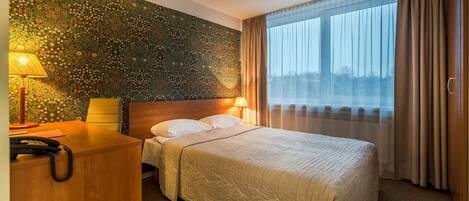 Quarto de hotel com cama grande, escrivaninha de madeira, telefone e janela com cortinas.