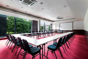 Un salón de conferencias con una larga mesa, sillas, una pantalla de proyector y ventanas amplias.