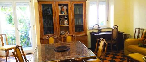 A dining area with a checkered floor, a wooden dining table, and chairs. There is a bookcase with books and decorative items.