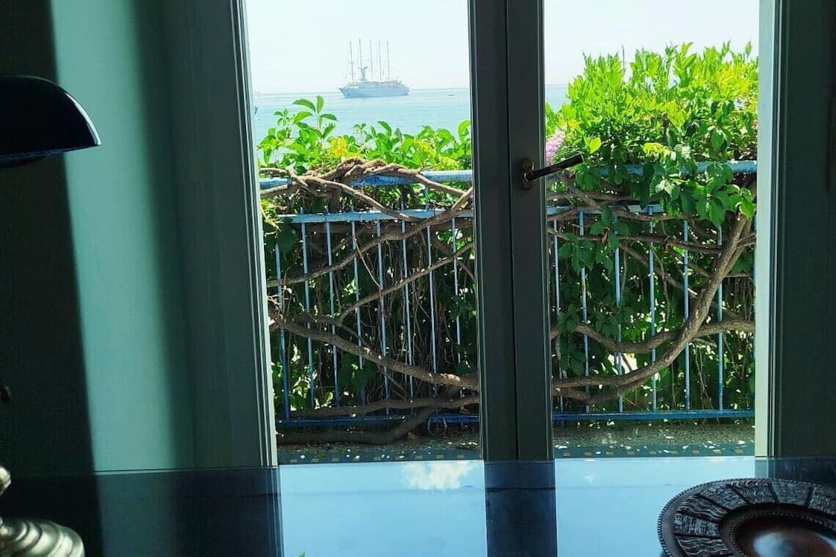 A room with a large window overlooking a balcony with lush greenery and a view of the sea.