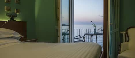 A hotel room with a bed, a chair, a small table with a vase, and a view of the sea through a balcony door.