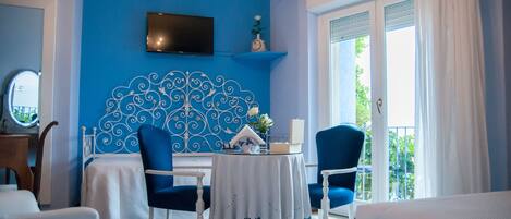A room with a blue accent wall, a flat-screen TV, a decorative headboard, a table with a white tablecloth, and two blue chairs.