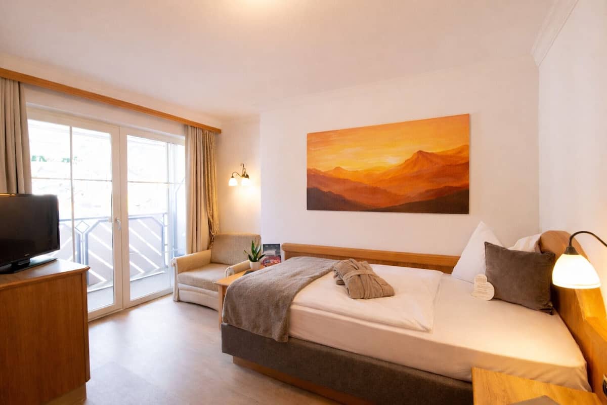 A hotel room with a bed, a chair, a desk with a TV, a painting on the wall, and a balcony with curtains.
