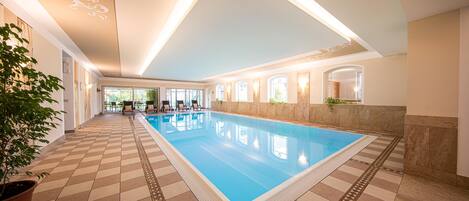 An indoor swimming pool with a tiled floor, a ceiling with decorative patterns, and a view of the outdoors through large windows.