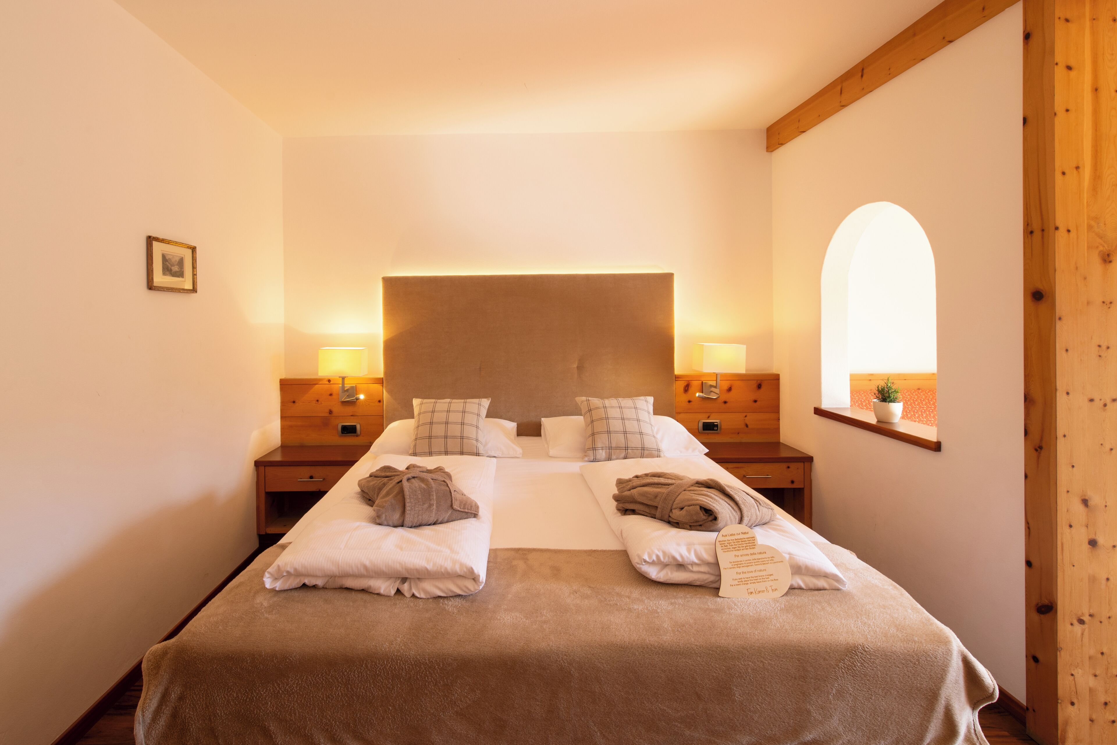 Una habitación de hotel con una cama grande, dos mesitas de noche, un cabecero, un ventanal con vista arqueada y una pequeña planta en maceta.