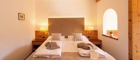 A hotel room with a large bed, two bedside tables, a headboard, a window with an arched view, and a small potted plant.