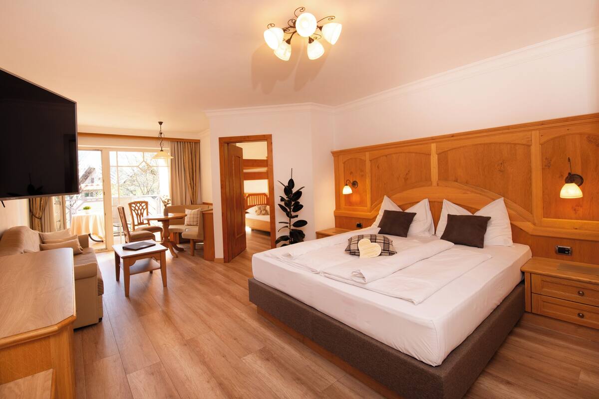 A hotel room with a large bed, a TV, a dining area, and a wooden floor.
