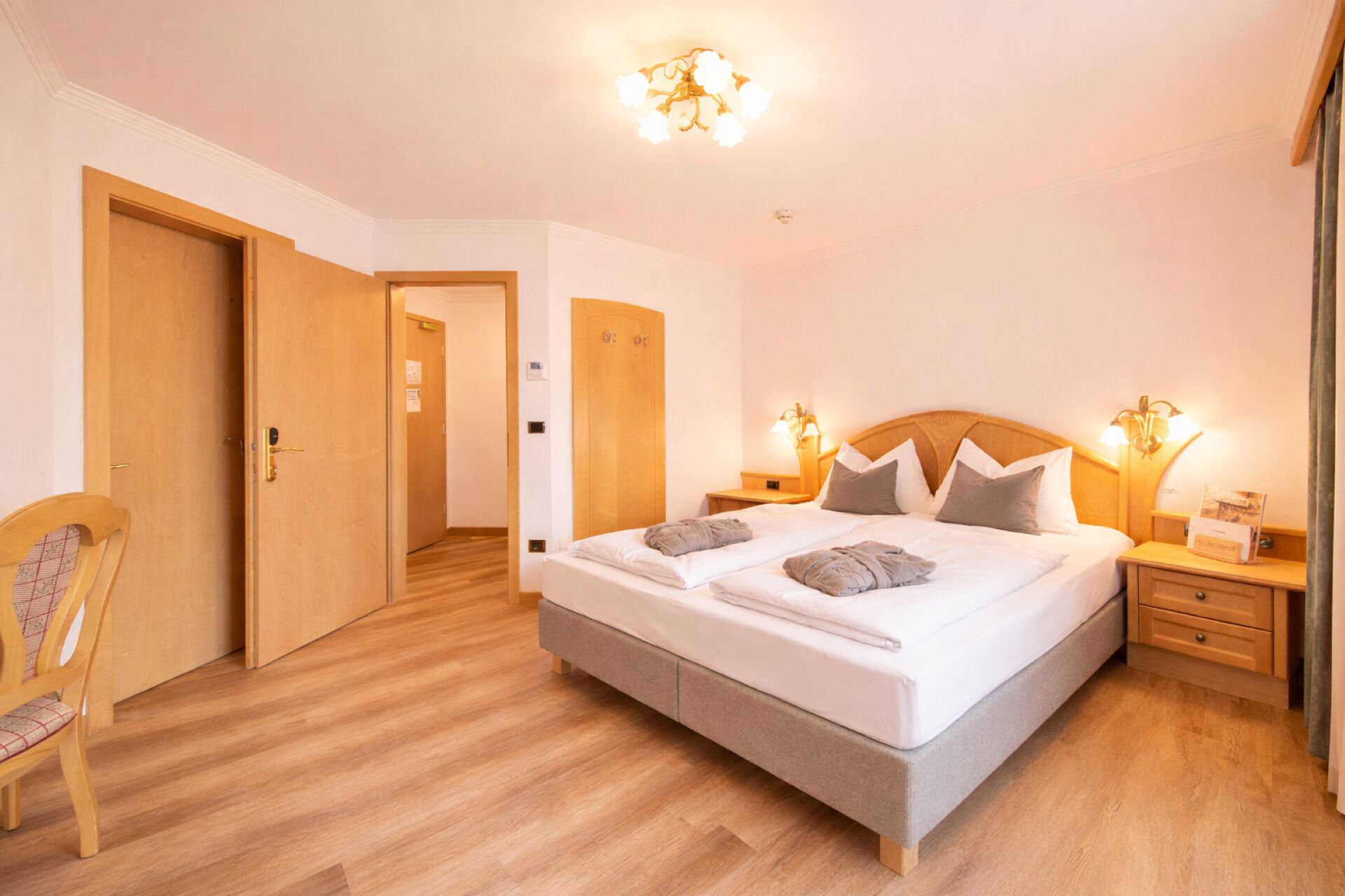 A hotel room with a large bed, wooden flooring, a chair, and a nightstand with a lamp.