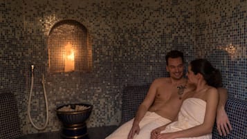 Una pareja sentada en un banco de azulejos azules en una habitación con paredes de mosaicos y una lámpara central.