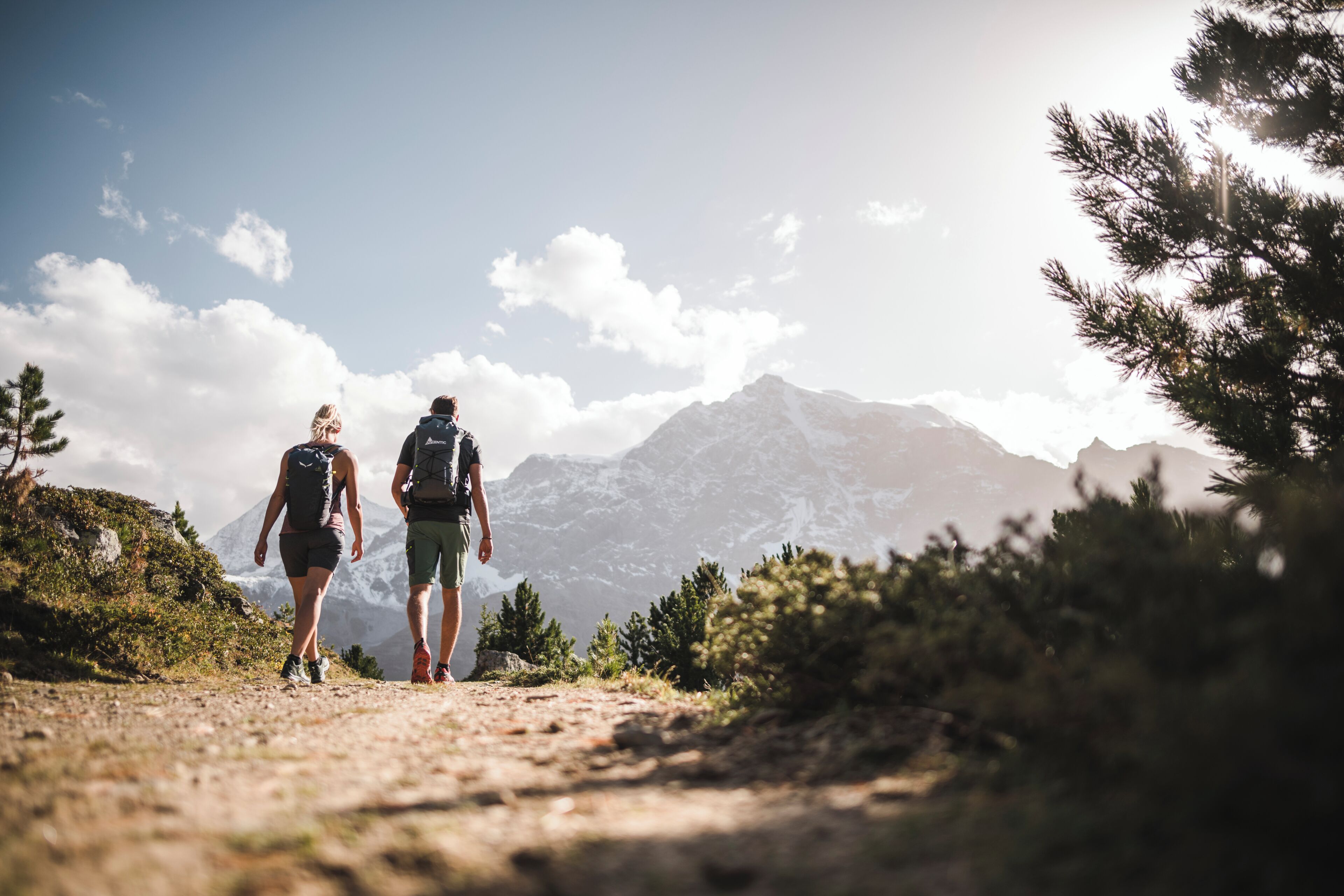 Zwei Personen wandern auf einem Bergpfad, im Hintergrund ein Berg.