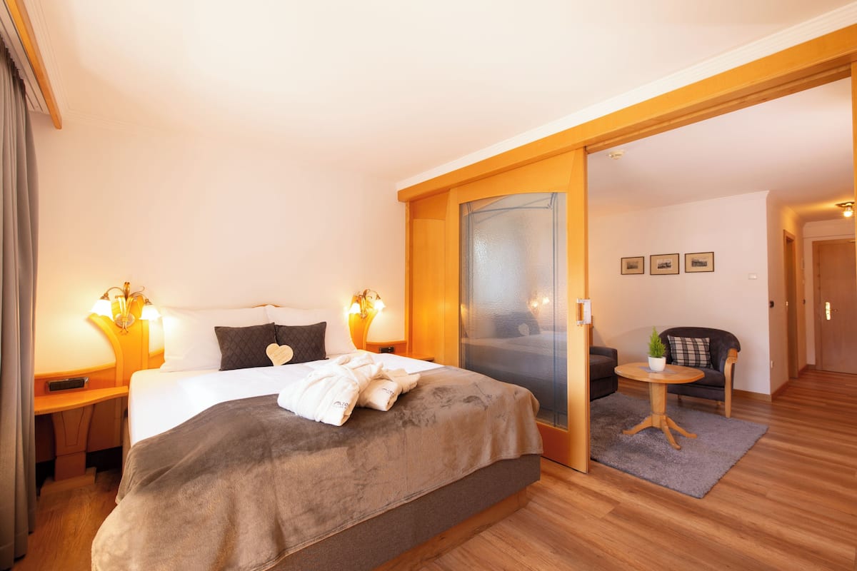 A hotel room with a large bed, bedside lamps, a small table with a plant, and a sofa in the adjacent living area.
