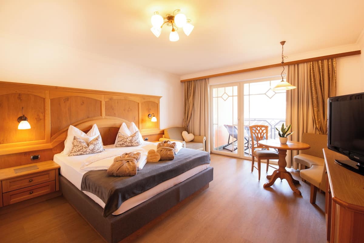 A hotel room with a bed, bedside tables, a TV, a dining area with a table and chairs, and a large window with curtains.