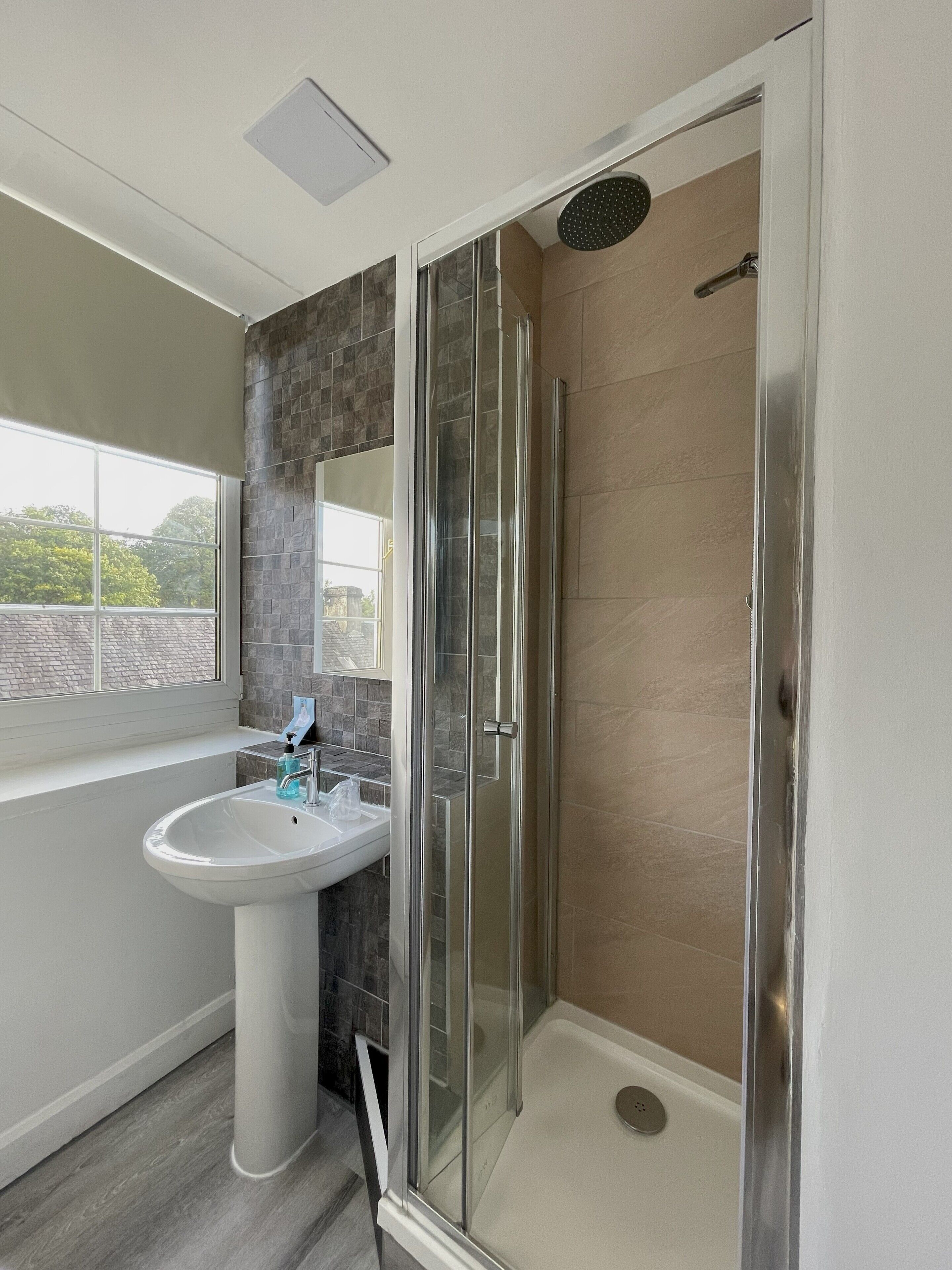 Un baño moderno con ducha envuelta en vidrio, lavamanos pedestal y ventana con vistas al exterior.