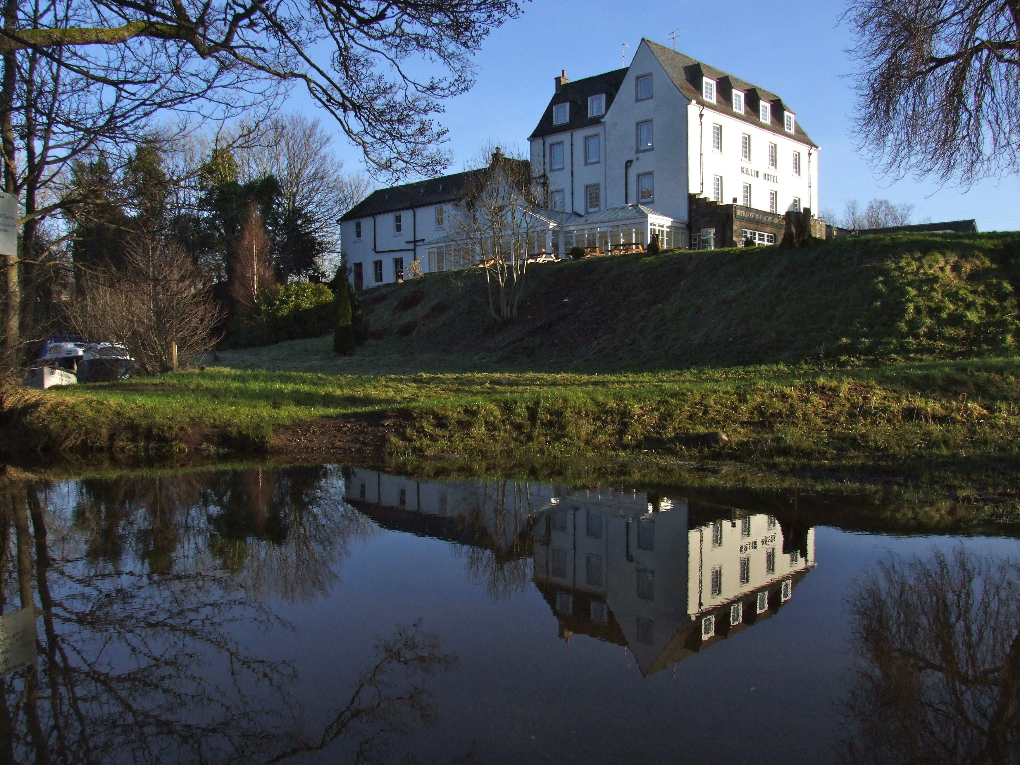 A large, white building with multiple windows and a sloped roof, reflecting in a calm body of water.