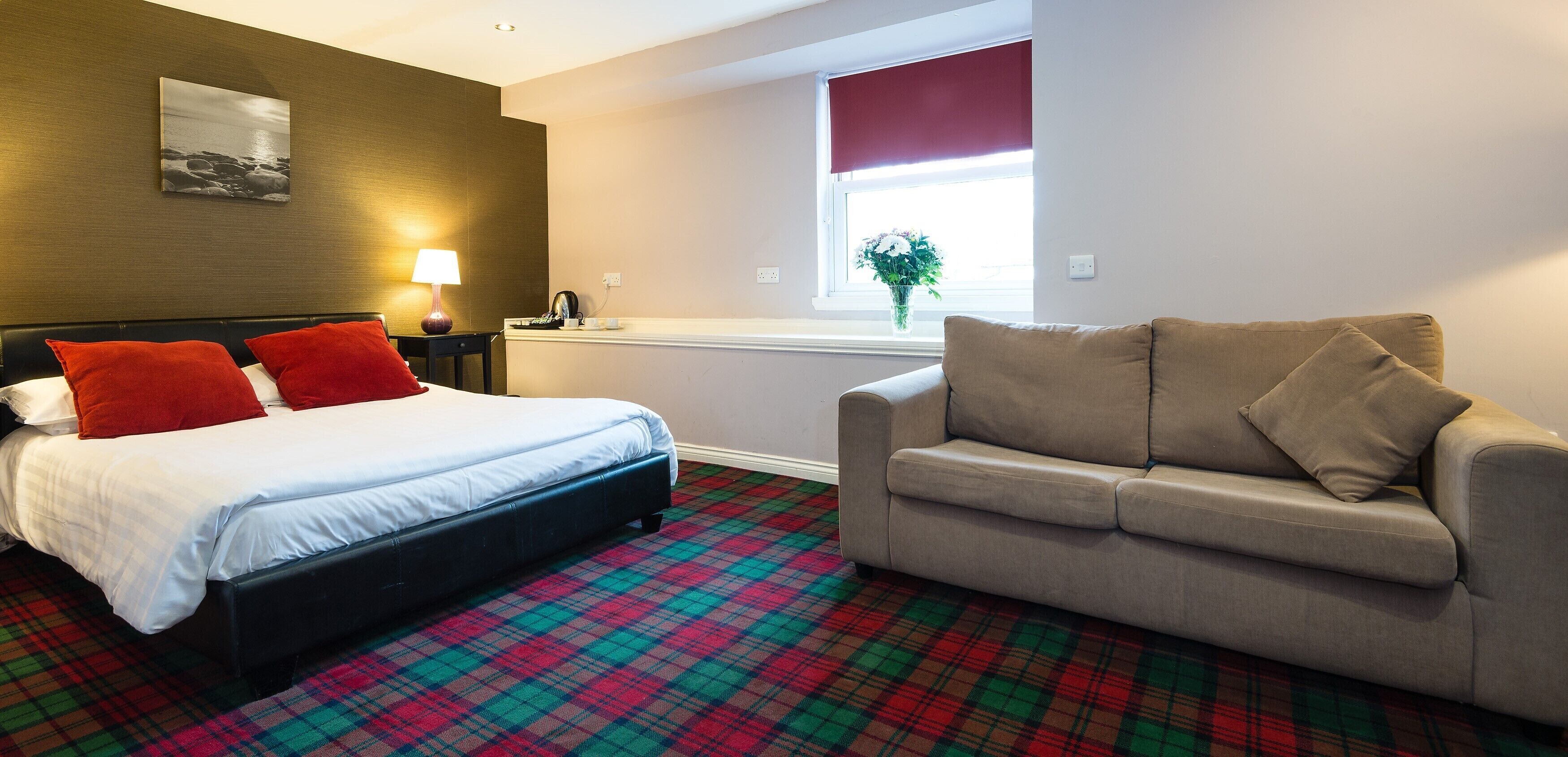A hotel room with a bed, a sofa, a plaid carpet, a window with a plant, and a wall-mounted picture.
