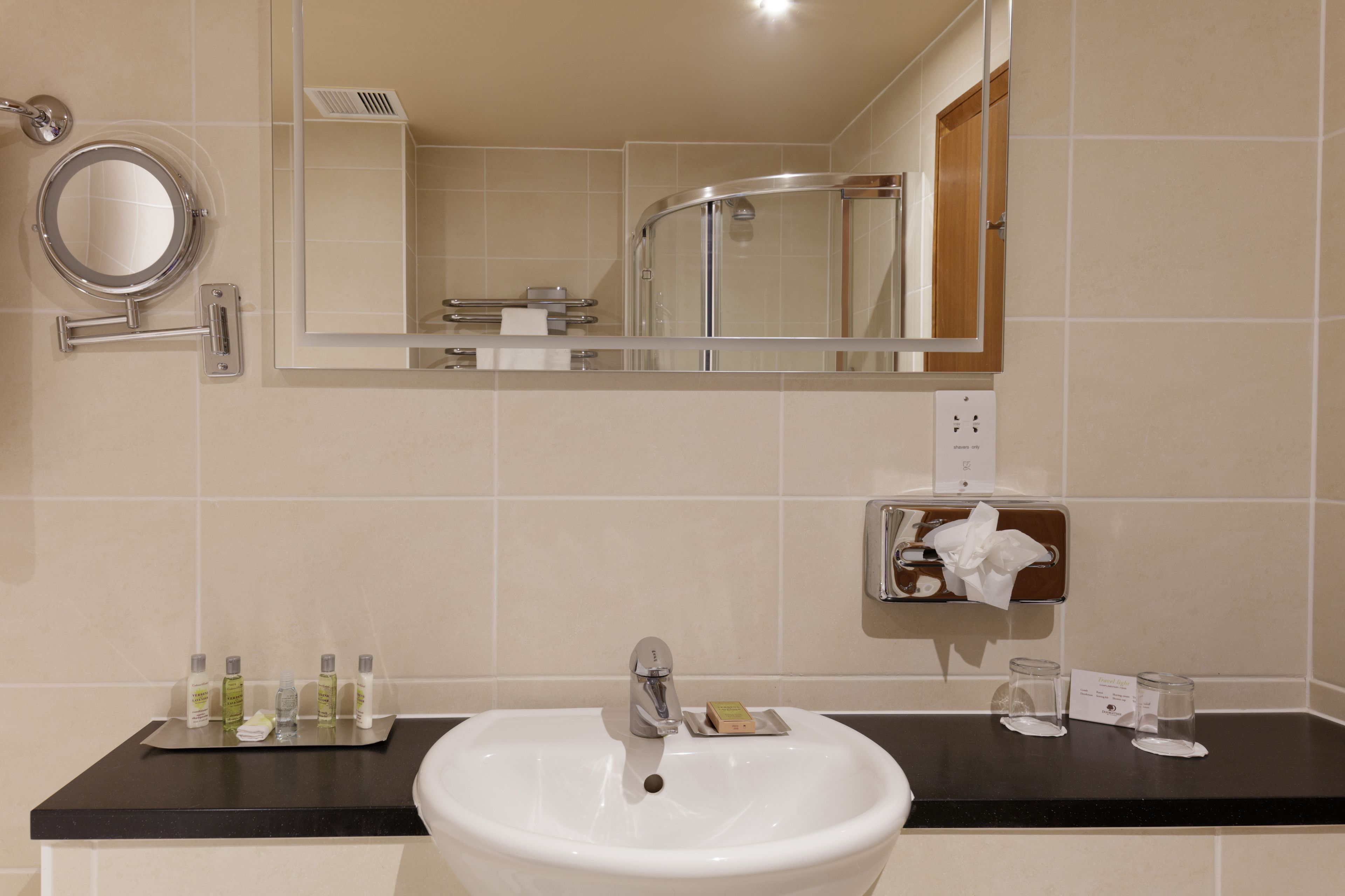 A bathroom with a white sink, a round mirror, a handheld mirror, a towel dispenser, and toiletries on the countertop.