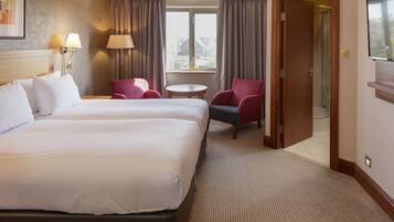 A hotel room with two beds, a sitting area with a table and chairs, a window with curtains, and a shower area.