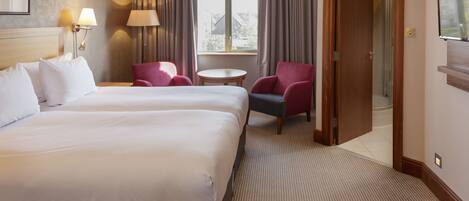 A hotel room with two beds, a sitting area with a table and chairs, a window with curtains, and a shower area.