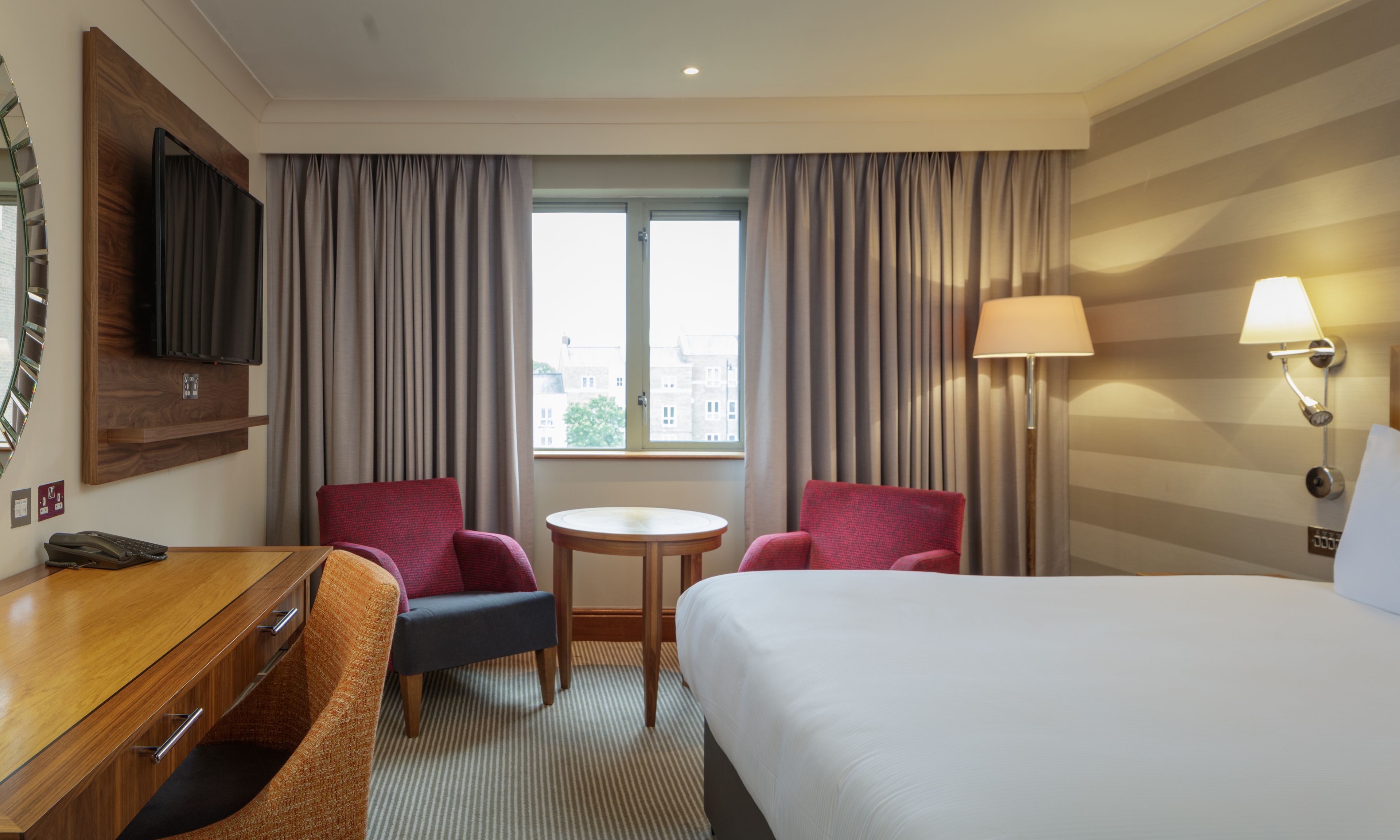 A hotel room with a bed, a desk with a telephone, two armchairs, a small round table, a lamp, and a window with curtains.
