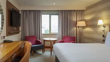 A hotel room with a bed, a desk with a telephone, two armchairs, a small round table, a lamp, and a window with curtains.