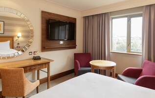 Habitación de hotel con cama, escritorio, silla, televisor montado en la pared, ventana con cortinas y vistas al exterior.