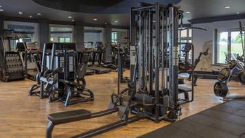 A gym with various exercise machines, a wooden floor, and large windows allowing natural light.