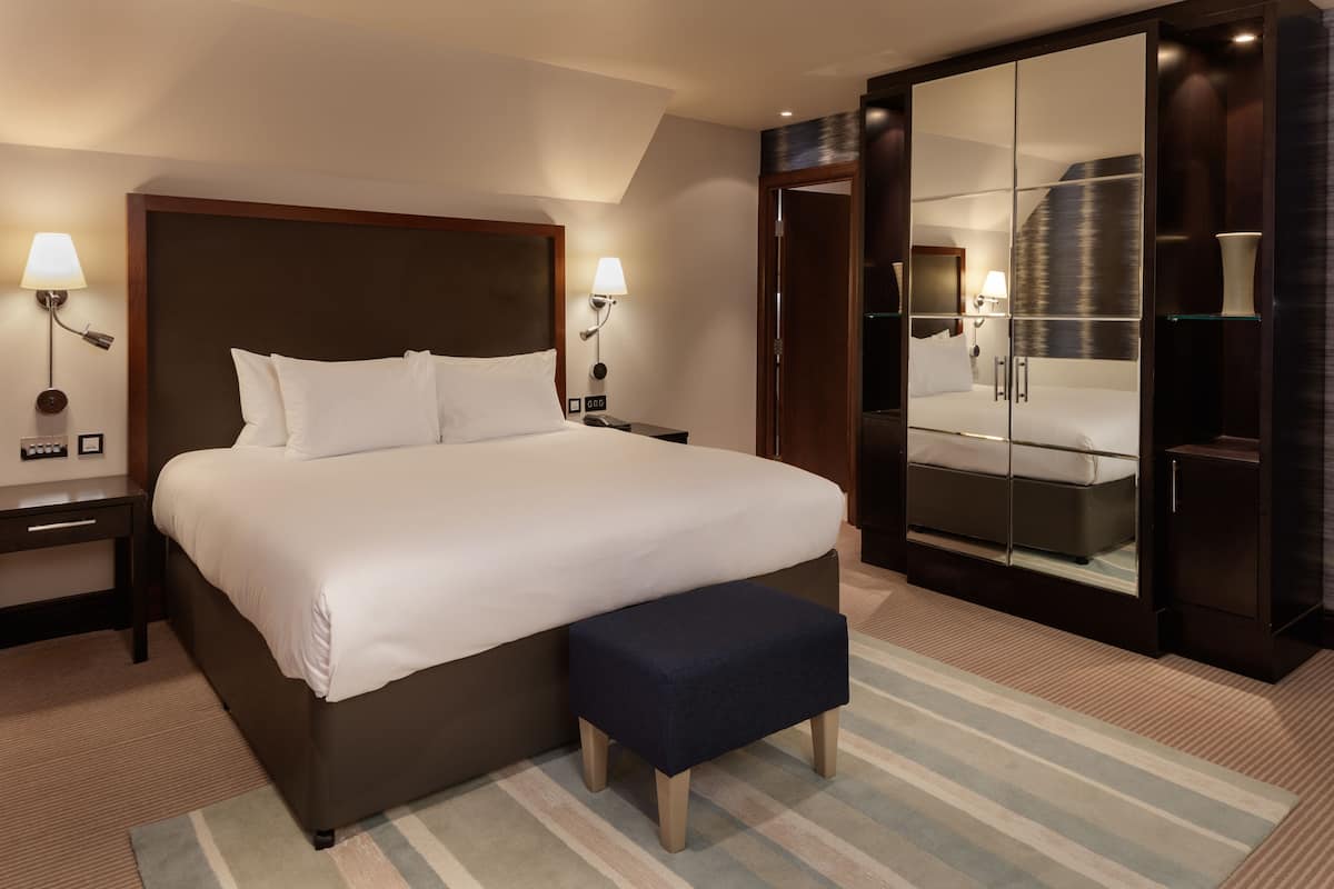 A hotel room with a large bed, bedside tables, a bench, a wardrobe, and a mirror.