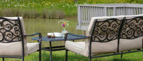 A table with chairs and a vase of flowers, set on a grassy area near a body of water and a wooden deck.