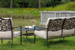 A table with chairs and a vase of flowers, set on a grassy area near a body of water and a wooden deck.