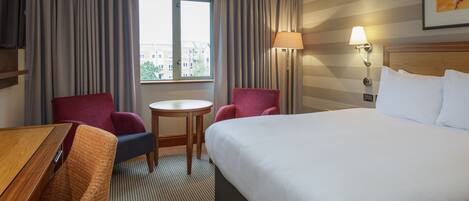A hotel room with a large bed, two armchairs, a small table, a TV, and a window with curtains.