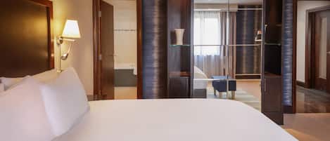 A hotel room with a large bed, a bedside lamp, a glass-enclosed bathroom, and a view of the city.