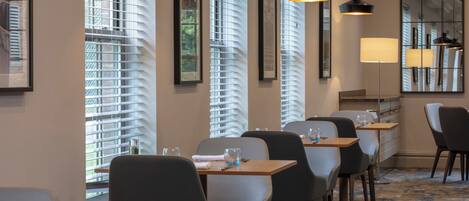Un espace de restauration d’hôtel avec des tables et des chaises, de grandes fenêtres équipées de stores et des luminaires suspendus.