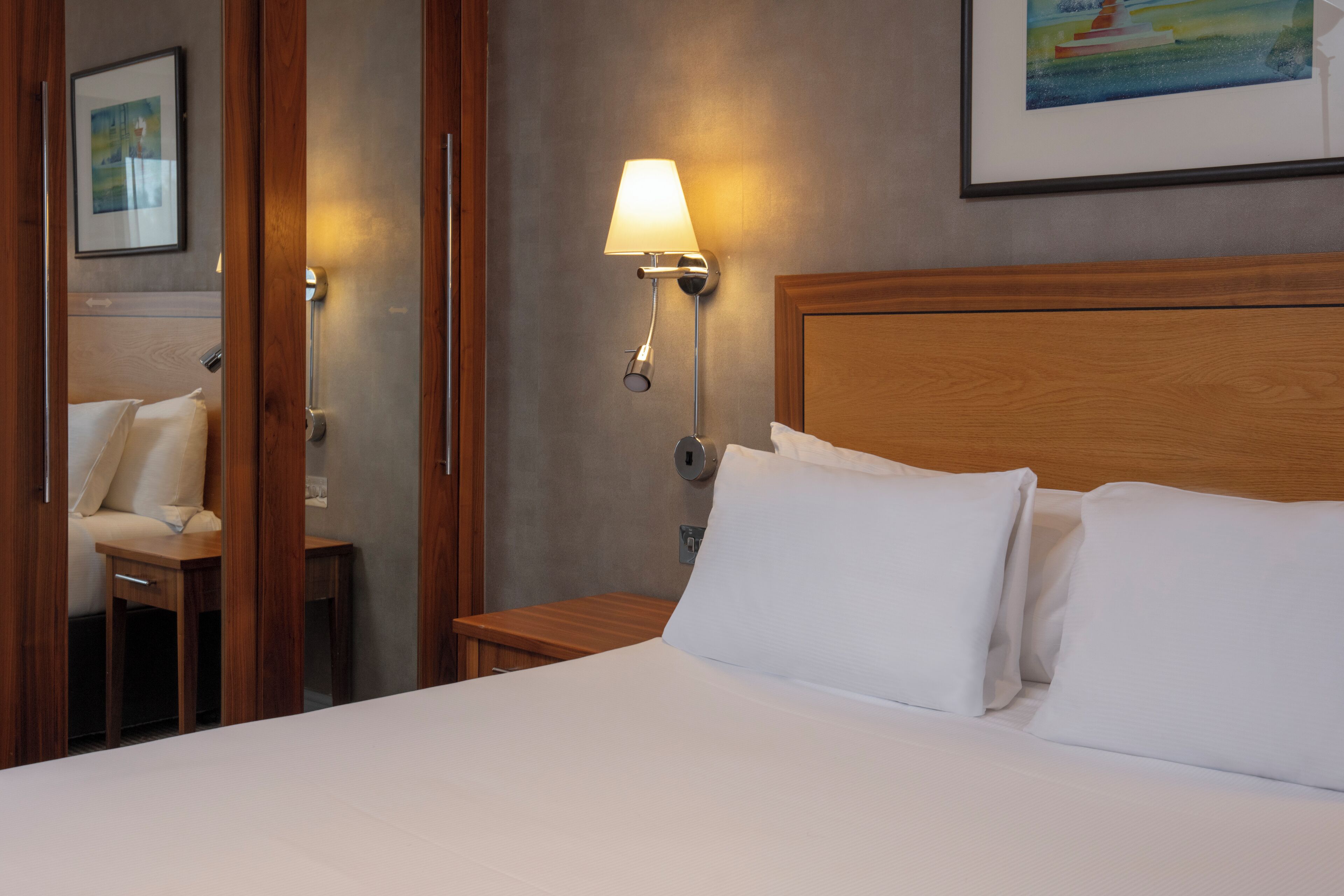 A hotel room with a neatly made bed, a bedside table with a lamp, and a painting on the wall.