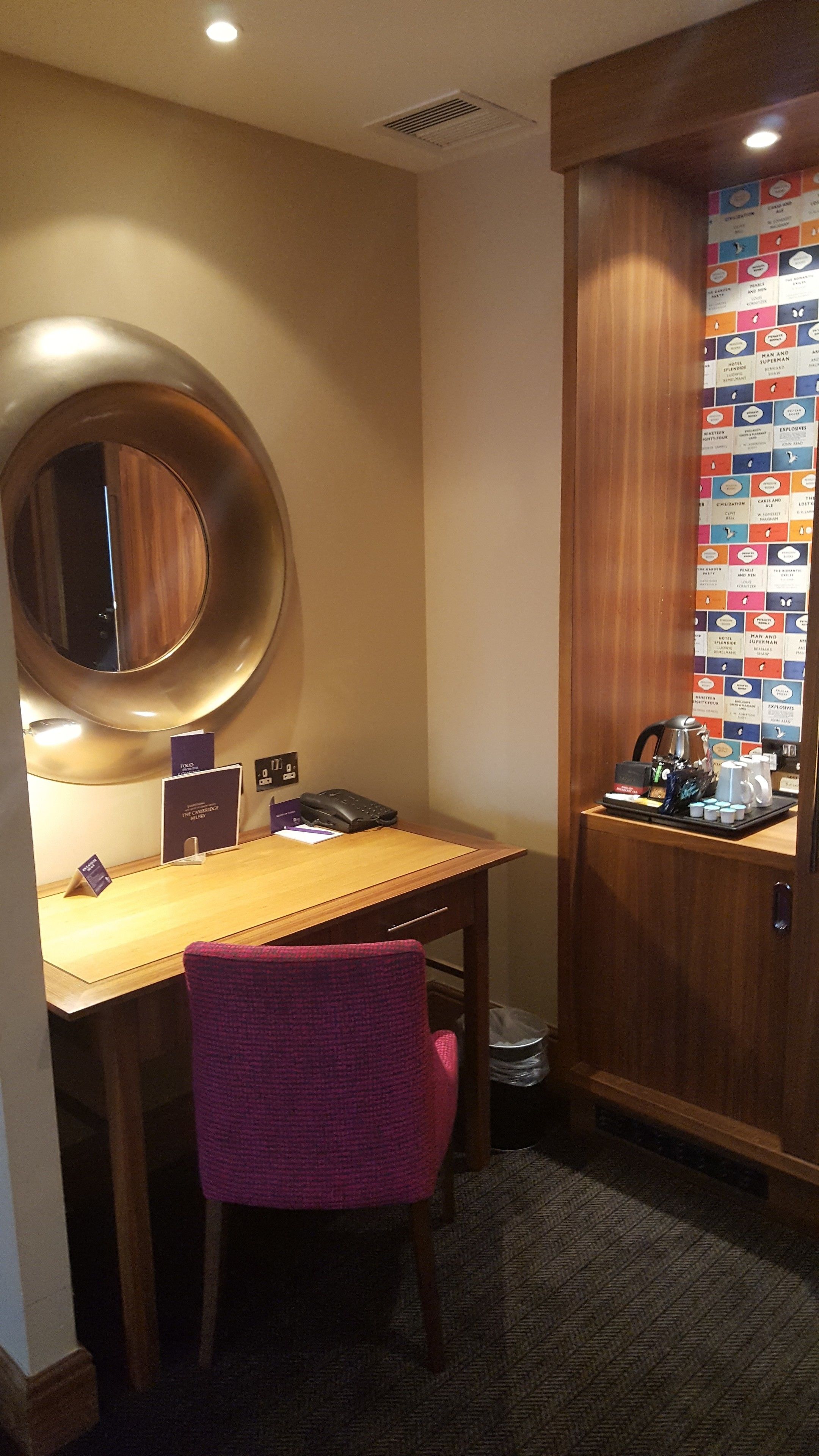 A hotel room with a wooden desk, a round mirror, a pink chair, and a wall with colorful book covers.