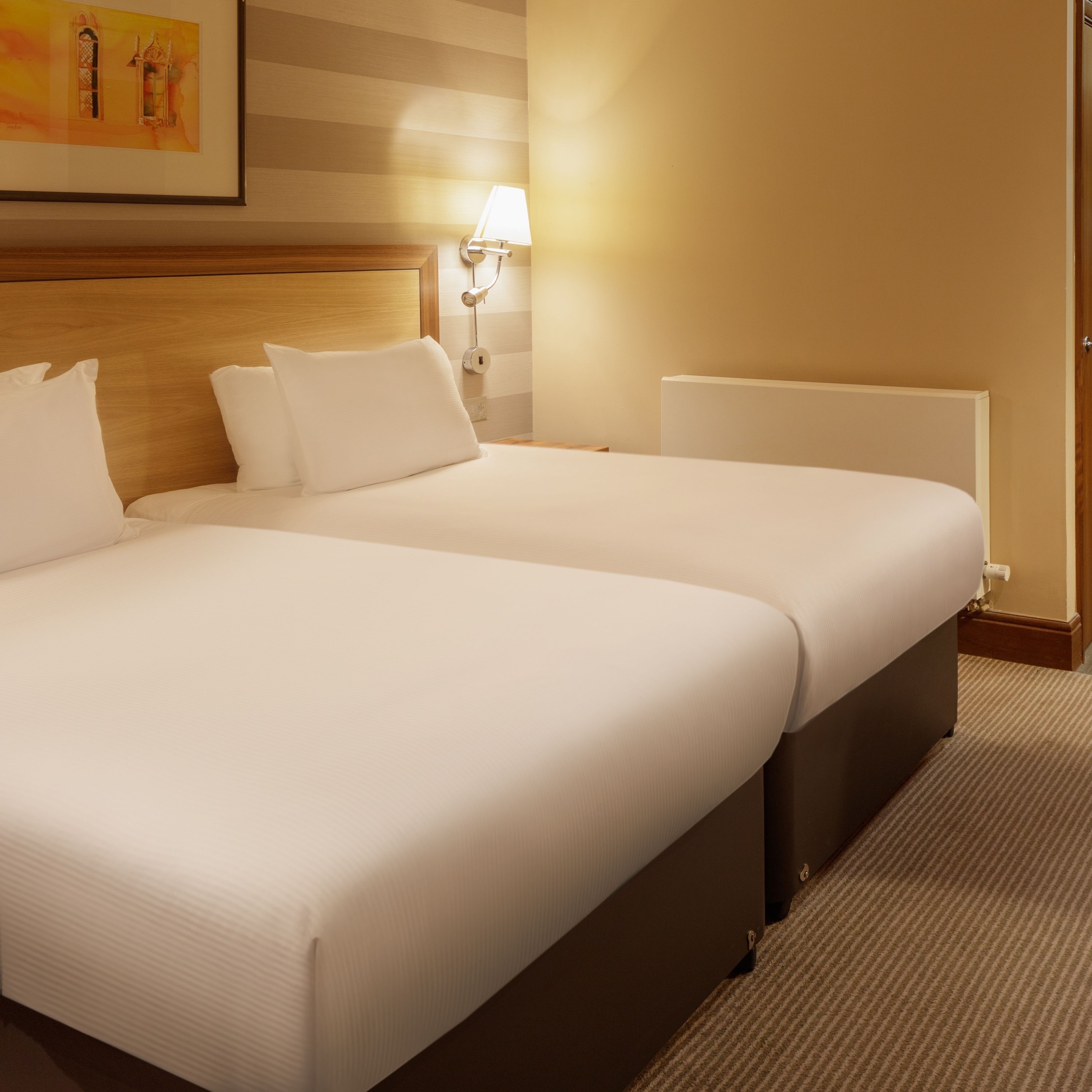 A hotel room with two beds, a nightstand with a lamp, a painting on the wall, and a radiator.