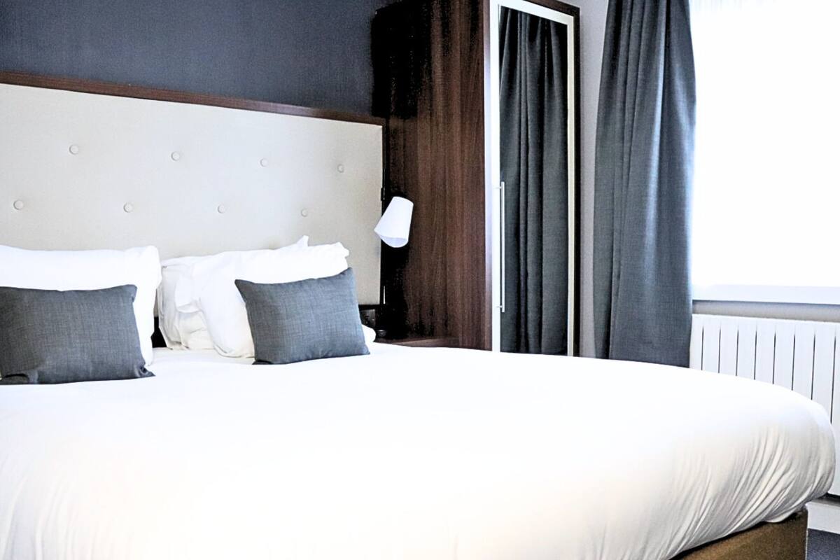 Habitación de hotel con una cama grande, dos almohadas, una mesita de noche, una lámpara y una ventana con cortinas.