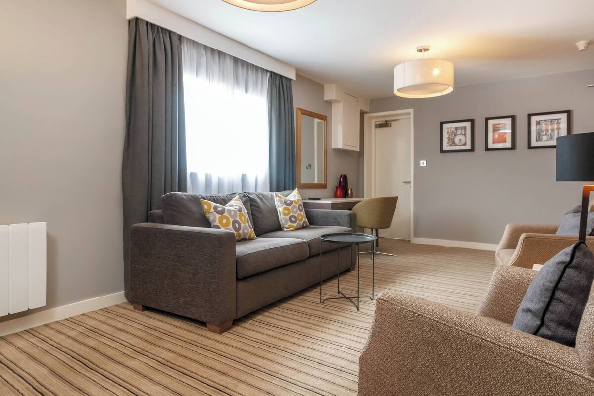 Una sala de estar moderna con un sofá gris, una mesita redonda pequeña y un sillón marrón. Hay un ventanal con cortinas, un espejo en la pared y cuadros enmarcados.