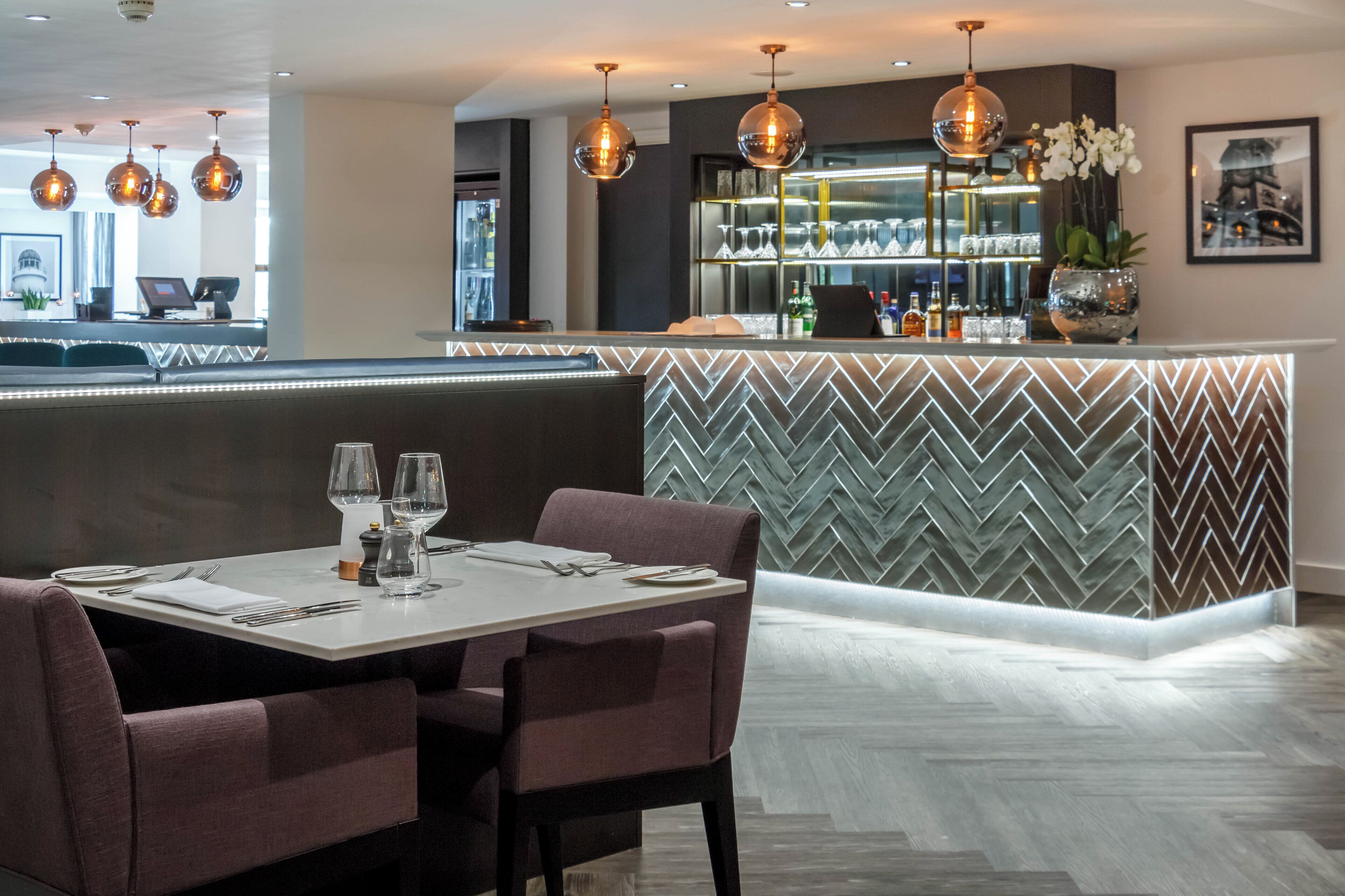 Een restaurant met een bar met een visgraatmotief, hanglampen en een zithoek met gestoffeerde zitzuilen.