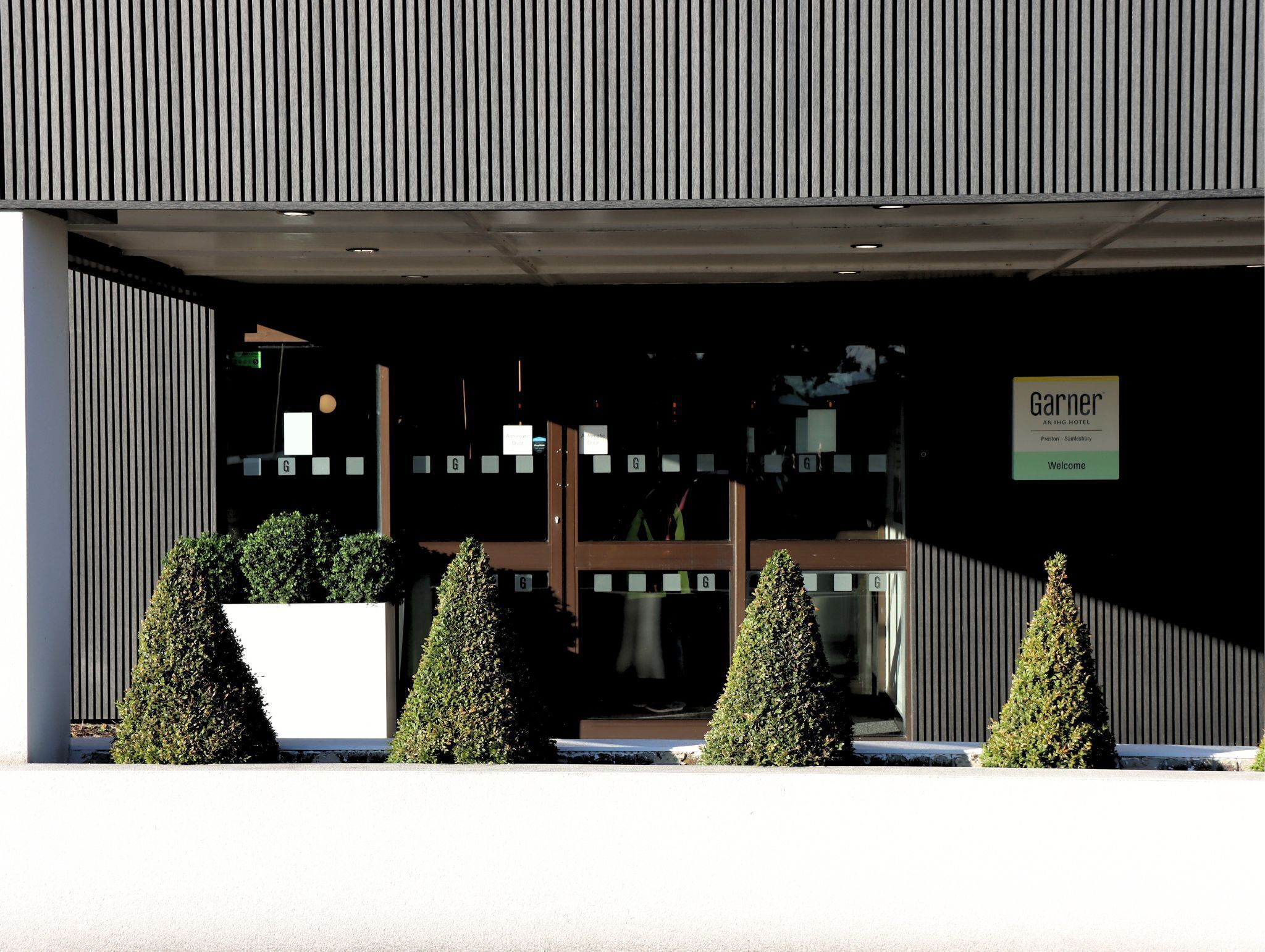 L’entrée d’un hôtel moderne, avec une façade sombre, une enseigne et des arbustes taillés avec soin.