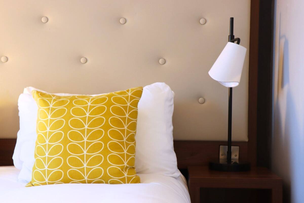 Una cama bien tendida con almohadillas blancas y amarillas estampadas, un cabecero capitonado y una mesita de noche con una lámpara.