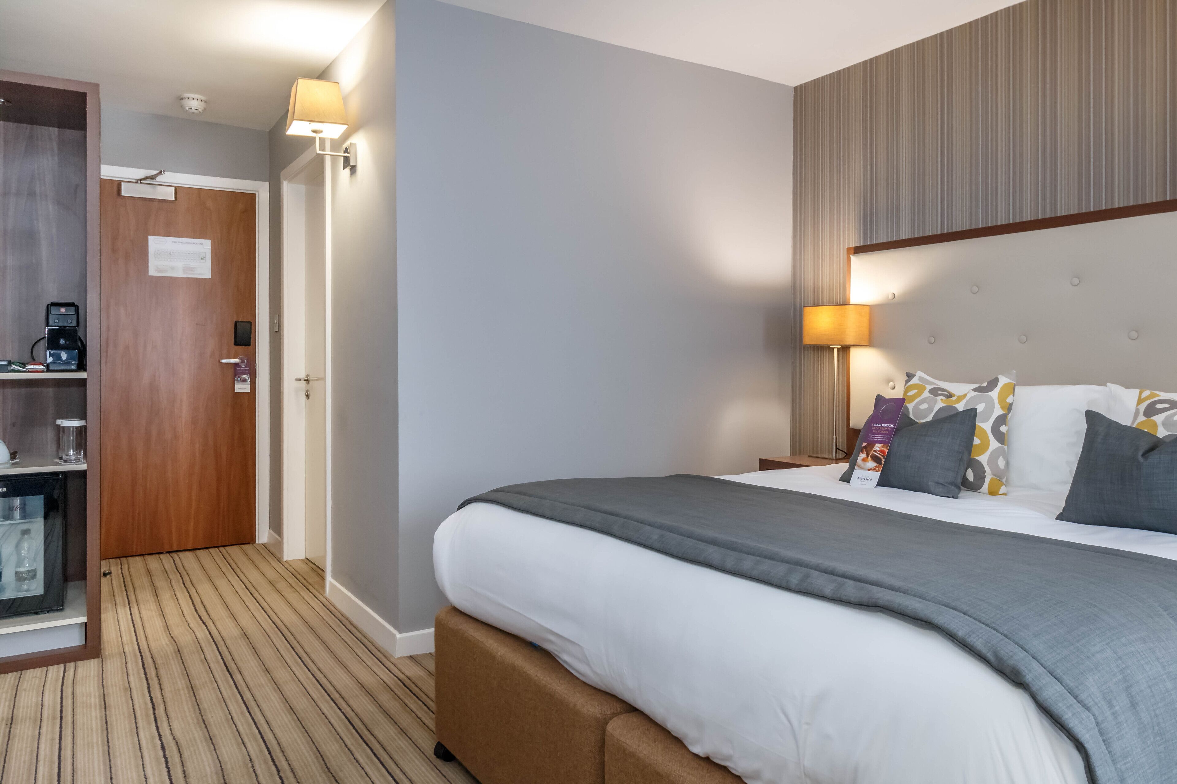 A hotel room with a large bed, a bedside table with a lamp, a minibar, and a door leading to a bathroom.