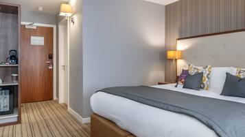 A hotel room with a large bed, a bedside table with a lamp, a minibar, and a door leading to a bathroom.