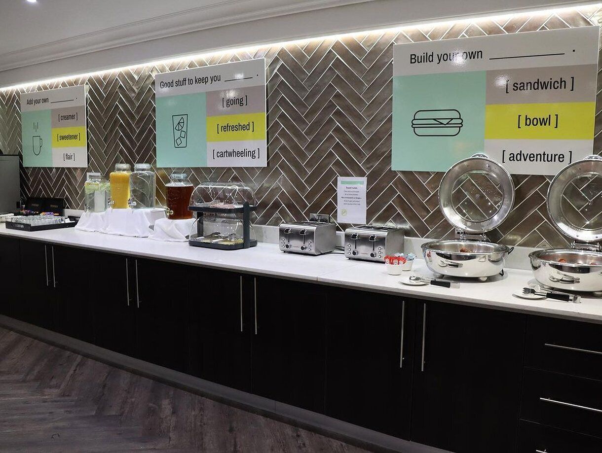 A buffet setup with various food items and utensils, including toasters and bowls, against a herringbone patterned wall.